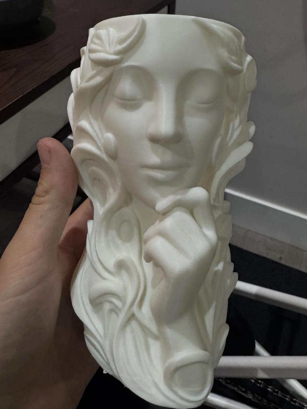 White 3D-printed vase sculpted as a woman's face with flowing hair and flowers, held in a hand