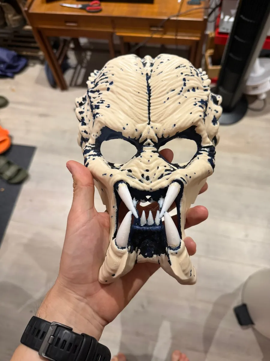Cream 3D-printed Predator mask with navy weathering spots and white tusks, held in a hand
